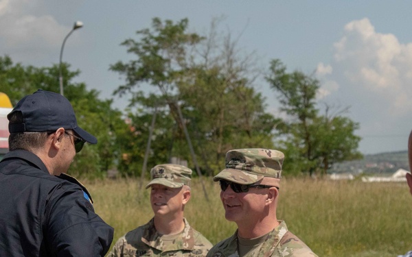 The image shows U.S. Army personnel in MultiCam camouflage uniforms engaged in a conversation with individuals in civilian and law enforcement attire outdoors. At least one soldier displays a 'KFOR' (Kosovo Force) patch and 'US ARMY' tape, indicating a NATO peacekeeping mission context. The setting appears to be an open area with vehicles, trees, and a partly cloudy sky in the background.