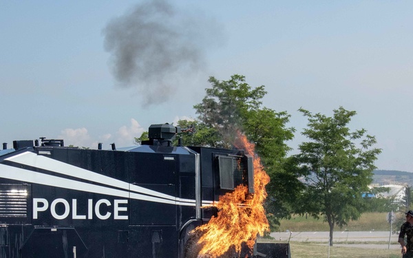 A large armored police vehicle is engulfed in flames on its right side, likely during a training exercise or demonstration. A person in camouflage clothing is visible to the right of the vehicle, appearing to move away from the fire. Yellow caution tape is strung in the foreground, suggesting a controlled perimeter around the event.