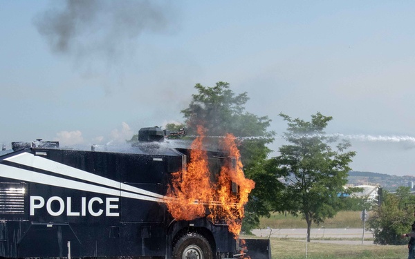 A large armored police vehicle is engulfed in flames on a paved surface, with fire visible around its midsection and undercarriage. At least two individuals are visible nearby, and yellow safety tape is strung in the foreground, suggesting a controlled demonstration or training exercise. A water stream is visible being discharged from the top of the vehicle.