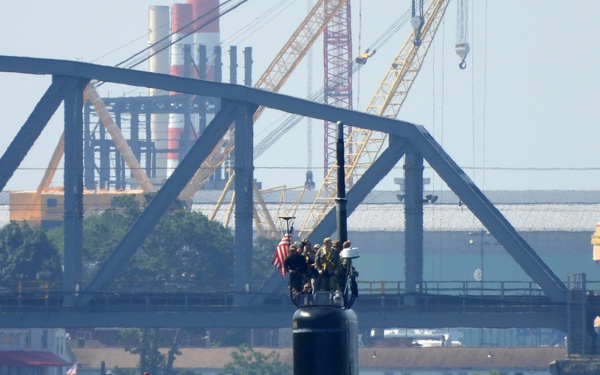 A U.S. Navy submarine is transiting through an urban waterway with crew members standing on the hull and sail (conning tower). An American flag is visible on the sail, and numerous sailors are positioned along the deck, suggesting a homecoming or departure ceremony. Industrial infrastructure including a steel truss bridge and large cranes are visible in the background.