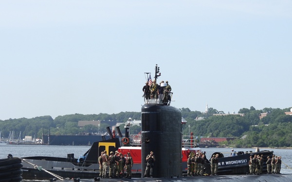 A large black military submarine is navigating a river or harbor, assisted by a red tugboat named 'N.P. Wronowski' visible on the port side. Numerous crew members in military/naval uniforms are stationed on the submarine's deck and atop the sail (conning tower), with an American flag visible at the top. The scene appears to be a homecoming or departure event at a naval facility.