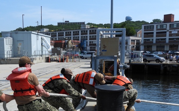 A group of Navy personnel in camouflage uniforms and orange life vests are working together to handle mooring lines at a pier, securing or releasing a vessel at a naval installation. Several sailors are pulling on a thick white rope around a large bollard on the dock edge. A waterfront facility with multi-story buildings and a police vehicle is visible in the background.