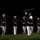 A nighttime military ceremonial drill performance featuring uniformed Marines in formal dress blues with white trousers and white covers. One Marine in the center is performing a rifle toss, with the rifle visibly airborne above the formation. All personnel are wearing black face masks, suggesting this was photographed during the COVID-19 pandemic era.