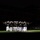 A nighttime military drill or ceremonial parade is taking place, with a group of uniformed personnel illuminated by a spotlight performing a rifle drill or manual of arms. The performers wear dark dress uniforms with white trousers, white gloves, and white covers (caps), consistent with U.S. Marine Corps dress blues. A second formation of similarly uniformed personnel stands at attention in the darker background to the right.