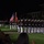 A formal military parade is taking place at night on a well-lit grass field, featuring uniformed Marines marching in precise formation. The ceremony includes a color guard carrying the United States flag and Marine Corps flags, with troops bearing rifles at shoulder arms. Spectators are visible in the foreground observing the event, and a brick building is visible in the background.