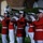 A military marching band, identifiable as the United States Marine Corps Band based on their distinctive red tunics and white trousers dress uniforms, performs at what appears to be an evening ceremonial event. Multiple musicians are playing large brass instruments, specifically mellophone or marching baritone-style horns, in a synchronized formation. The setting appears to be an outdoor venue with stone architecture and manicured hedges visible in the background.