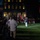 A nighttime military ceremony is taking place on a well-lit lawn in front of a large illuminated building. Marines in dress uniforms with red tunics and white trousers are assembled in formation, with a color guard prominently displaying the American flag and other flags. A crowd of civilian spectators observes the ceremony from the left side.