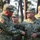 Two U.S. Army soldiers in OCP (Operational Camouflage Pattern) uniforms are shaking hands in what appears to be an outdoor military ceremony or formal greeting. The soldier on the right has a name tape reading 'MOORE' and a 'U.S. ARMY' tape, with a single star insignia visible indicating a Brigadier General rank. Several other uniformed soldiers are visible in the background among trees.