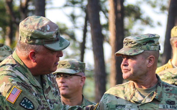 Two U.S. Army soldiers in OCP (Operational Camouflage Pattern) uniforms are shaking hands in what appears to be an outdoor military ceremony or formal greeting. The soldier on the right has a name tape reading 'MOORE' and a 'U.S. ARMY' tape, with a single star insignia visible indicating a Brigadier General rank. Several other uniformed soldiers are visible in the background among trees.