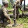 Two U.S. Army soldiers in MultiCam/OCP camouflage uniforms are setting up a mortar system in a wooded training area. The foreground soldier is kneeling and steadying the mortar tube while a second soldier handles associated equipment. Additional soldiers are visible in the background, suggesting a unit-level field training exercise.