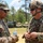 Two U.S. Army soldiers in MultiCam (OCP) uniforms are engaged in conversation in an outdoor field environment. The soldier on the left wears a patrol cap and has 'AIRBORNE' and a 25th Infantry Division patch visible, while the soldier on the right wears a combat helmet and has the name tape 'WOODRUFF' visible. Military vehicles including HMMWVs and a tactical truck are visible in the background.