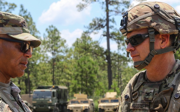 Two U.S. Army soldiers in MultiCam (OCP) uniforms are engaged in conversation in an outdoor field environment. The soldier on the left wears a patrol cap and has 'AIRBORNE' and a 25th Infantry Division patch visible, while the soldier on the right wears a combat helmet and has the name tape 'WOODRUFF' visible. Military vehicles including HMMWVs and a tactical truck are visible in the background.
