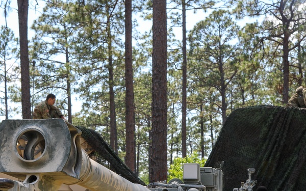 The image shows military personnel operating what appears to be an M198 155mm howitzer in a wooded training area. Soldiers are working around the artillery piece, which is positioned near a military truck, with camouflage netting deployed over nearby equipment. The setting appears to be a field exercise or training operation in a pine forest environment.