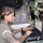 A female soldier in U.S. Army ACU (Army Combat Uniform) is working at a laptop computer inside what appears to be a military communications or command vehicle. She is focused on the screen while typing, surrounded by rack-mounted electronic equipment, cables, and military gear. The setting suggests a field operations or signals/communications environment.