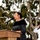 A woman in a black dress speaks at a wooden podium with a microphone at what appears to be an outdoor military ceremony. In the background, a naval officer in a white dress uniform is rendering a salute. The setting includes trees and an overcast sky, suggesting a formal commemorative or official event.