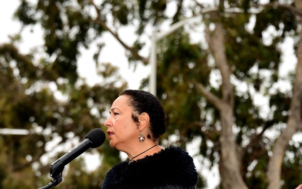 A woman in a black dress speaks at a wooden podium with a microphone at what appears to be an outdoor military ceremony. In the background, a naval officer in a white dress uniform is rendering a salute. The setting includes trees and an overcast sky, suggesting a formal commemorative or official event.