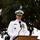 A senior military officer in a white dress uniform is speaking at a podium with a microphone, smiling during what appears to be an official ceremony or formal event held outdoors. The officer is wearing a white combination cover with gold insignia, multiple service ribbons, medals, and gold shoulder boards indicating high rank. The setting features trees in the background under an overcast sky.
