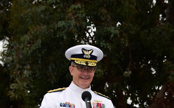 A senior military officer in a white dress uniform is speaking at a podium with a microphone, smiling during what appears to be an official ceremony or formal event held outdoors. The officer is wearing a white combination cover with gold insignia, multiple service ribbons, medals, and gold shoulder boards indicating high rank. The setting features trees in the background under an overcast sky.