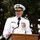 A senior military officer in white dress uniform is speaking at a podium with a microphone, likely at an official ceremony or formal event held outdoors. The uniform features extensive medal ribbons, gold insignia, and shoulder boards indicating high rank. The setting appears to be a daytime outdoor event with trees visible in the background.