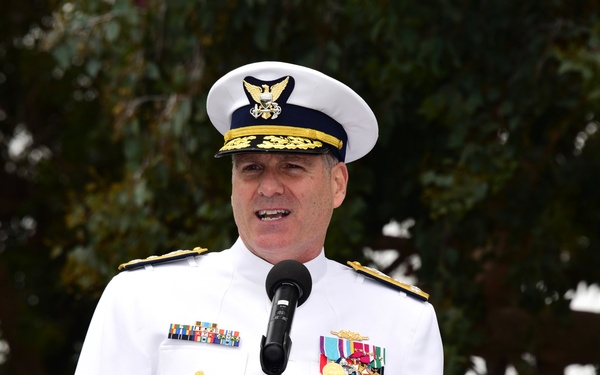 A senior military officer in white dress uniform is speaking at a podium with a microphone, likely at an official ceremony or formal event held outdoors. The uniform features extensive medal ribbons, gold insignia, and shoulder boards indicating high rank. The setting appears to be a daytime outdoor event with trees visible in the background.