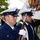 Two U.S. Coast Guard members in dress uniform are participating in a formal color guard ceremony outdoors. They are standing at attention in profile view, with one member holding a ceremonial rifle. A colorful flag or guidon with streamers is visible in the background, along with a gathered crowd suggesting an official military event.
