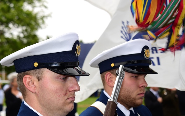 Two U.S. Coast Guard members in dress uniform are participating in a formal color guard ceremony outdoors. They are standing at attention in profile view, with one member holding a ceremonial rifle. A colorful flag or guidon with streamers is visible in the background, along with a gathered crowd suggesting an official military event.