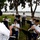 A military band performs outdoors near a waterfront marina, with musicians seated in a semicircle facing a conductor. The performers are dressed in white dress uniforms with black berets and blue trousers, consistent with U.S. Army dress attire. The setting appears to be a park or ceremonial outdoor venue with trees, grass, and boats visible in the background.