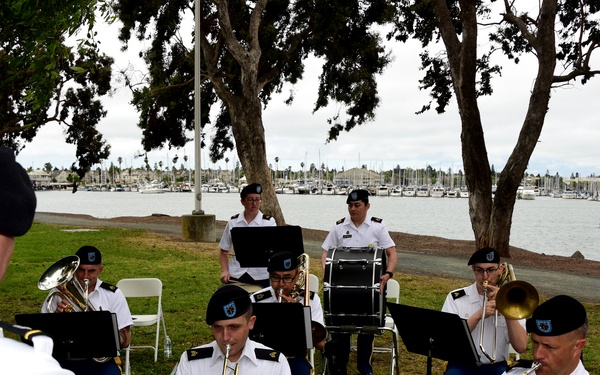 A military band performs outdoors near a waterfront marina, with musicians seated in a semicircle facing a conductor. The performers are dressed in white dress uniforms with black berets and blue trousers, consistent with U.S. Army dress attire. The setting appears to be a park or ceremonial outdoor venue with trees, grass, and boats visible in the background.
