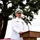 A senior military officer in white dress uniform stands at a wooden podium delivering a speech at an outdoor ceremony. The officer wears a white combination cover with gold insignia, multiple service ribbons, and medals, indicating high rank. The setting appears to be a formal military event held outdoors beneath a large tree.