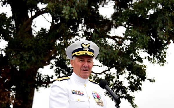 A senior military officer in white dress uniform stands at a wooden podium delivering a speech at an outdoor ceremony. The officer wears a white combination cover with gold insignia, multiple service ribbons, and medals, indicating high rank. The setting appears to be a formal military event held outdoors beneath a large tree.