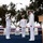 A formal United States Coast Guard change of command or awards ceremony is taking place outdoors on a blue-carpeted stage. Two officers in white dress uniforms are exchanging salutes while a third officer stands between them as a presiding official. The setting includes patriotic red, white, and blue bunting decorations and a Coast Guard seal-emblazoned podium.