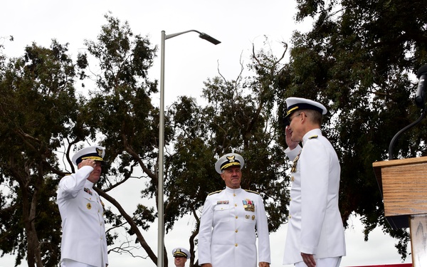 A formal United States Coast Guard change of command or awards ceremony is taking place outdoors on a blue-carpeted stage. Two officers in white dress uniforms are exchanging salutes while a third officer stands between them as a presiding official. The setting includes patriotic red, white, and blue bunting decorations and a Coast Guard seal-emblazoned podium.