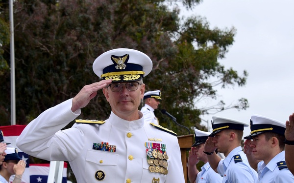 A senior military officer in a white dress uniform with extensive medal decorations is rendering a salute during a formal outdoor ceremony. He is flanked by rows of junior personnel in light blue shirts and navy trousers who are also saluting. The setting appears to be an official change of command or military honors ceremony.