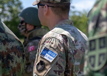 The image shows a group of multinational military personnel standing together, likely during a joint NATO operation or exercise. The central figure wears a NATO KFOR (Kosovo Force) RC-EAST patch on their MultiCam uniform, indicating participation in the Kosovo peacekeeping mission. Multiple nations appear to be represented based on the different camouflage patterns visible.