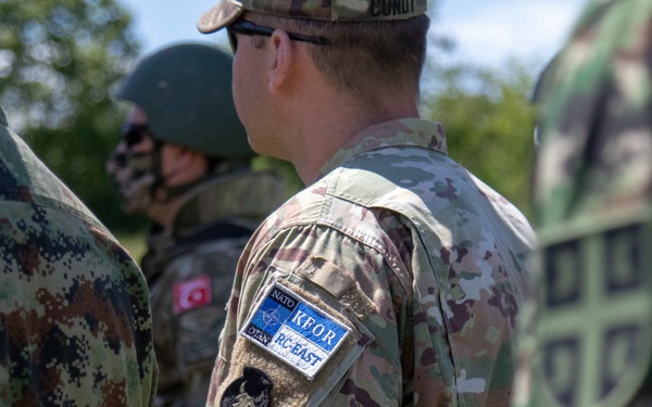 The image shows a group of multinational military personnel standing together, likely during a joint NATO operation or exercise. The central figure wears a NATO KFOR (Kosovo Force) RC-EAST patch on their MultiCam uniform, indicating participation in the Kosovo peacekeeping mission. Multiple nations appear to be represented based on the different camouflage patterns visible.