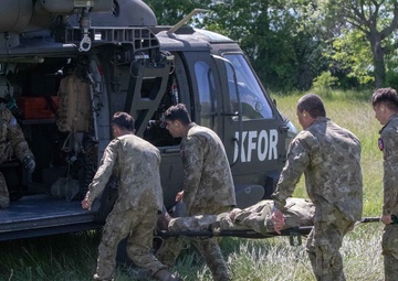Military personnel in camouflage uniforms are loading or unloading a casualty on a stretcher into/from a military helicopter marked 'KFOR' in a grassy field. Four soldiers are carrying the stretcher while at least one crew member is visible inside the helicopter. This appears to be either a medical evacuation (MEDEVAC) operation or a training exercise conducted by KFOR (Kosovo Force) personnel.