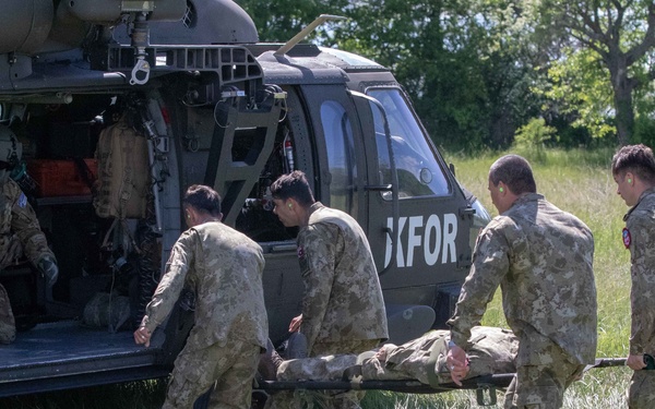 Military personnel in camouflage uniforms are loading or unloading a casualty on a stretcher into/from a military helicopter marked 'KFOR' in a grassy field. Four soldiers are carrying the stretcher while at least one crew member is visible inside the helicopter. This appears to be either a medical evacuation (MEDEVAC) operation or a training exercise conducted by KFOR (Kosovo Force) personnel.