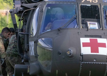 A military medical evacuation (MEDEVAC) helicopter, identifiable by the prominent Red Cross symbol on its fuselage, is landed in a grassy field. Several soldiers in camouflage uniforms are gathered at the side door of the aircraft, appearing to load or unload personnel or equipment. At least two crew members are visible in the cockpit.