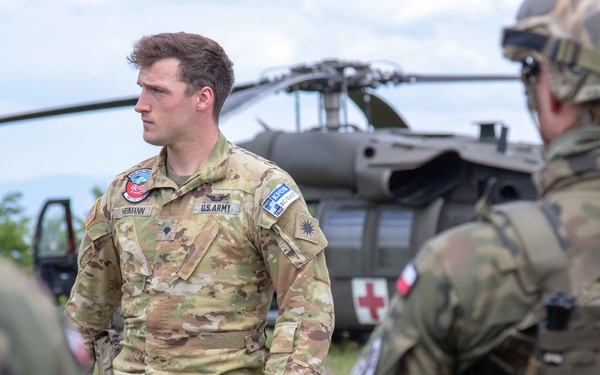 A U.S. Army soldier, identified by name tape as 'Hermann' and wearing OCP (Operational Camouflage Pattern) uniform, stands in a briefing or coordination group with other military personnel. A medical evacuation (MEDEVAC) helicopter marked with a red cross is visible in the background. The scene appears to be a multinational military exercise or operation, with soldiers wearing different national insignia present.