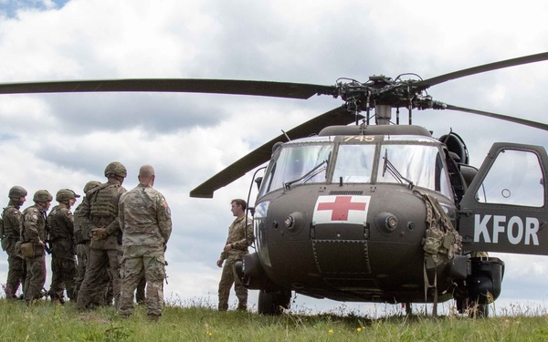 A military medical evacuation (MEDEVAC) helicopter, marked with a Red Cross and 'KFOR' insignia, is landed on a grassy field. A group of soldiers in camouflage uniforms are gathered near the aircraft, appearing to conduct a briefing or casualty transfer operation. The scene is consistent with a NATO Kosovo Force (KFOR) medical training exercise or operational activity.