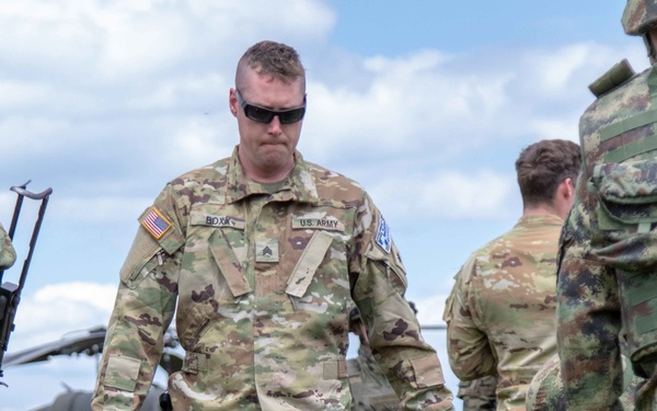 A U.S. Army Sergeant identified by the name tape 'BOXX' stands in a grassy field during what appears to be a joint military training exercise. He is wearing OCP (Operational Camouflage Pattern) uniform with a U.S. flag patch and a unit patch on the left shoulder, and is looking down at a simulated casualty on a litter. Other soldiers in different camouflage patterns are visible in the background near a military helicopter.