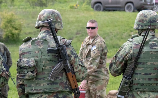 The image shows a group of military personnel from at least two different forces gathered in an outdoor grassy area, appearing to be engaged in a briefing or coordination activity. Several soldiers wearing distinctive pixelated camouflage uniforms and helmets carry AK-pattern rifles slung on their backs, while others in MultiCam/OCP uniforms appear to be instructors or liaison personnel. The setting suggests a joint military training exercise or field coordination meeting.