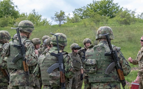 A group of soldiers in camouflage uniforms are gathered in an outdoor field setting, appearing to receive a briefing or instruction from a soldier in MultiCam/OCP pattern uniform. Several soldiers are carrying AK-pattern rifles on their backs along with tactical plate carriers. The setting appears to be a military training exercise or joint operation, with green rolling terrain in the background.