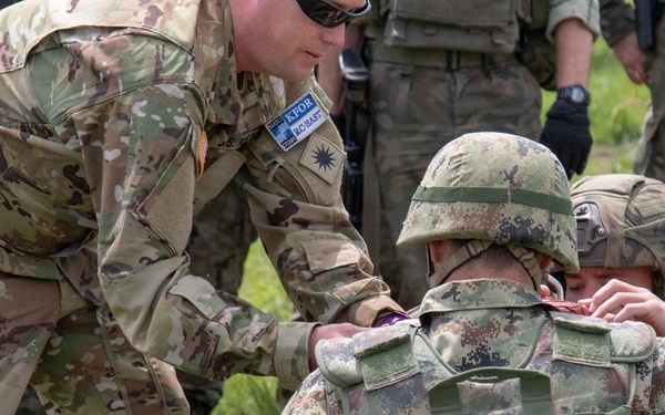 A U.S. Army soldier in MultiCam/OCP uniform is assisting or instructing a soldier wearing a different camouflage pattern (likely Eastern European or Balkan digital camo) during what appears to be a multinational military training exercise. The standing soldier has visible NATO KFOR (Kosovo Force) and RC-East patches on his uniform. Multiple other military personnel are visible in the background observing the activity.