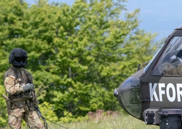 A fully equipped military personnel in camouflage uniform and black flight helmet stands in a grassy field beside a dark military helicopter marked 'KFOR' (Kosovo Force - NATO-led peacekeeping mission). The soldier appears to be connected to the helicopter via a cable or rope, suggesting a rappelling, fast-rope, or ground crew operation. The setting is an open field with dense green trees in the background under a partly cloudy sky.