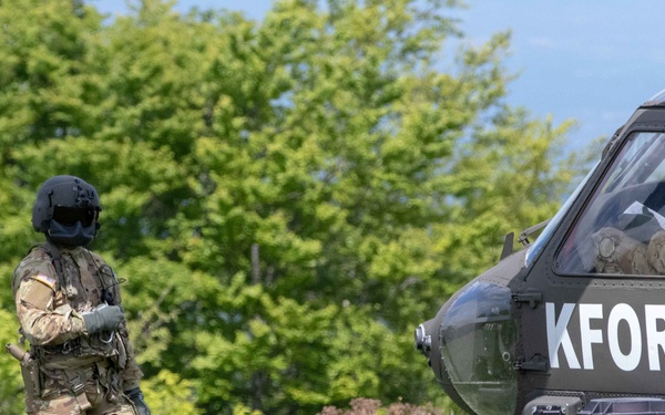 A fully equipped military personnel in camouflage uniform and black flight helmet stands in a grassy field beside a dark military helicopter marked 'KFOR' (Kosovo Force - NATO-led peacekeeping mission). The soldier appears to be connected to the helicopter via a cable or rope, suggesting a rappelling, fast-rope, or ground crew operation. The setting is an open field with dense green trees in the background under a partly cloudy sky.
