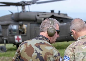 Two military personnel in camouflage uniforms are standing in a field, viewed from behind, appearing to converse while observing a UH-60 Black Hawk helicopter marked with KFOR insignia and a red cross medical symbol. The helicopter is on or near the ground in the background. Patches visible on one uniform indicate KFOR (Kosovo Force) and MNB-EAST (Multinational Battle Group East) affiliation.