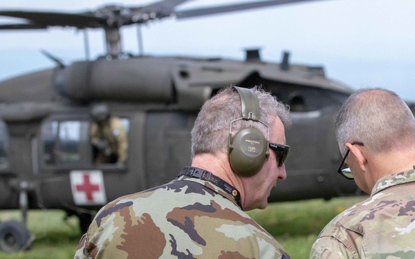 Two military personnel in camouflage uniforms are standing in a field, viewed from behind, appearing to converse while observing a UH-60 Black Hawk helicopter marked with KFOR insignia and a red cross medical symbol. The helicopter is on or near the ground in the background. Patches visible on one uniform indicate KFOR (Kosovo Force) and MNB-EAST (Multinational Battle Group East) affiliation.