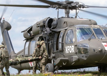 A military medical evacuation (MEDEVAC) operation is depicted, showing soldiers carrying a casualty on a stretcher toward a UH-60 Black Hawk helicopter marked with KFOR insignia and a Red Cross symbol. The helicopter, numbered 745, is landed on an open grassy field with wildflowers. This appears to be either a real MEDEVAC operation or a training exercise conducted under NATO's Kosovo Force (KFOR) mission.