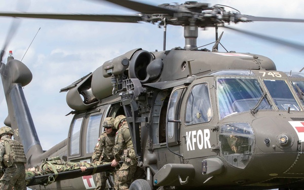 A military medical evacuation (MEDEVAC) operation is depicted, showing soldiers carrying a casualty on a stretcher toward a UH-60 Black Hawk helicopter marked with KFOR insignia and a Red Cross symbol. The helicopter, numbered 745, is landed on an open grassy field with wildflowers. This appears to be either a real MEDEVAC operation or a training exercise conducted under NATO's Kosovo Force (KFOR) mission.